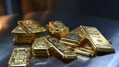 صورة تظهر سبائك ذهبية متراكمة مع خلفية لسوق الأسهم العالمية، تعكس ارتفاع أسعار الذهب وسط التوترات الجيوسياسية وتقلبات الأسواق المالية.