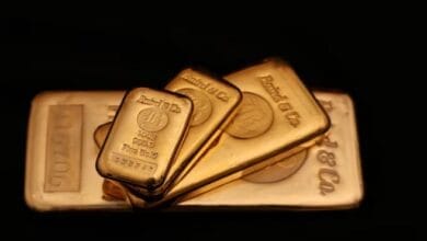 صورة لسبائك ذهبية متراكمة بجانب رسوم بيانية تظهر انخفاض الأسعار، مع خلفية تحمل عملات دولار أمريكي، ترمز لتأثر أسعار الذهب بالسياسات النقدية وتقلبات السوق.