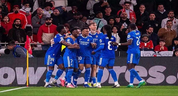 صورة للاعب فينيسيوس جونيور وهو يحتفل بهدفه في مرمى بنفيكا، مع إبراز شعاري ريال مدريد ودوري أبطال أوروبا في الخلفية.