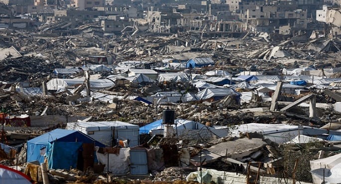 صورة جوية لمخيمات النازحين في قطاع غزة خلال شهر رمضان، تظهر خياماً متلاصقة بين ركام المباني المدمرة، مع أطفال يتجمعون حول نقطة توزيع مساعدات إنسانية من الأونروا.