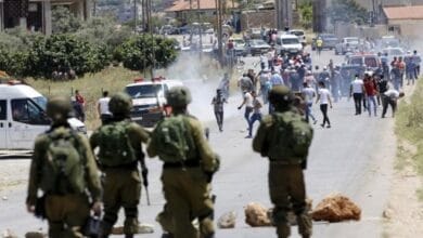 صورة لمواجهات عنيفة بين شبان فلسطينيين وقوات الاحتلال أو المستوطنين في الضفة الغربية، مع تصاعد دخان قنابل الغاز في الخلفية، تعكس أحداث العنف في عين يبرود.