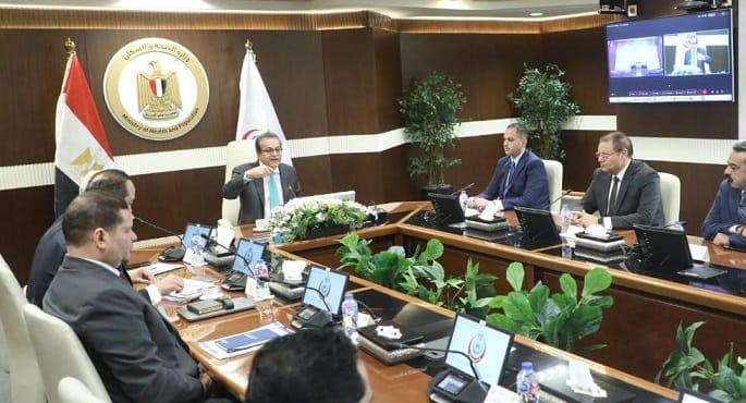 صورة لاجتماع رسمي رفيع المستوى يضم معالي وزير الصحة والسكان ومحافظ المنيا ومسؤولين آخرين، يناقشون خطط وإجراءات التشغيل التجريبي لمنظومة التأمين الصحي الشامل في محافظة المنيا، مع التركيز على الوثائق والشاشات التوضيحية.